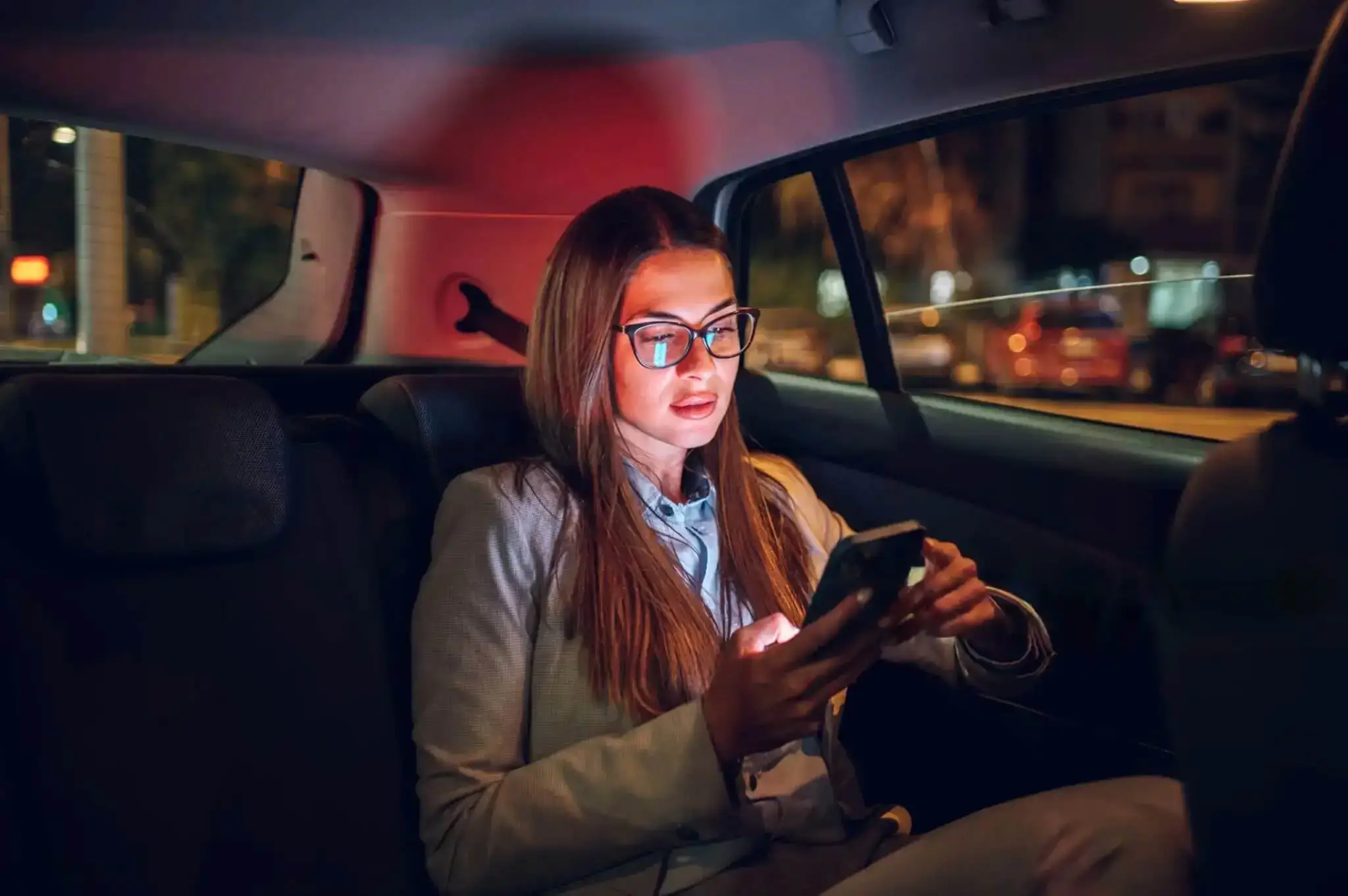 Business traveler sitting in the back seat of a car at night, using her phone during a Corsicana taxi ride to DFW Airport, representing reliable Corsicana Taxi service.